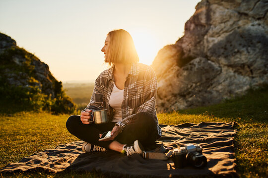 Young woman relaxing outdoors at golden hour with coffee mug and camera beside rocky hillside