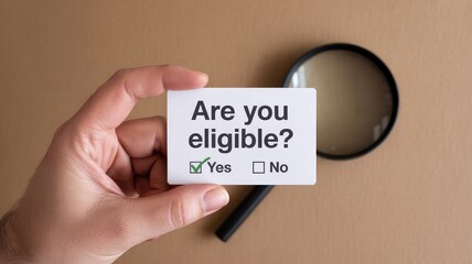 Person holding up a small card asking are you eligible with a magnifying glass nearby on desk