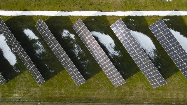 Top down drone view slowly descending above rows of solar panels generating renewable energy with spring snow patches visible on green grass.