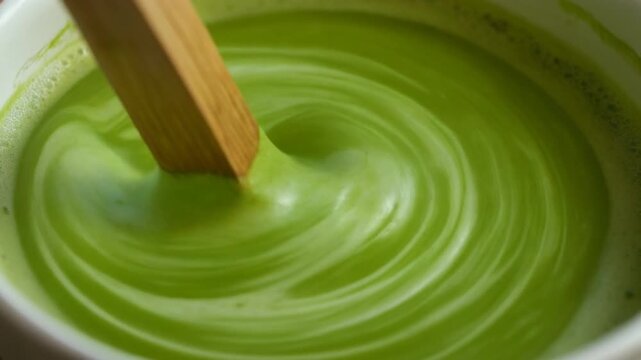 Mixing vibrant green matcha in a bowl with a wooden spoon, creating a swirling pattern from above.