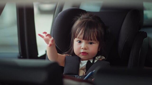 Happy little Asian girl sitting safely in car seat and looking out the window, toddler secured in child safety seat for road trip, concept of family travel, passenger safety.