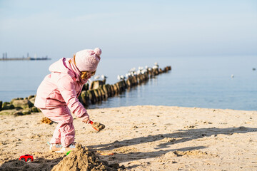 Child girl wearing warm winter clothes playing with sand on beach. Toddler using pink toy shovel....