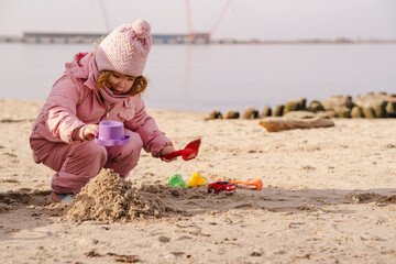 Child girl wearing warm winter clothes playing with sand on beach. Toddler using pink toy shovel....