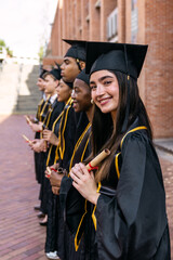 Group of happy multiracial graduates smiling, wearing academic gowns and caps, holding diplomas after successful university education