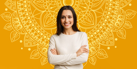 A woman stands with her arms crossed, smiling. Behind her is a large mandala design in bright yellow. The setting conveys warmth, and the atmosphere appears lively and cheerful. © Prostock-studio