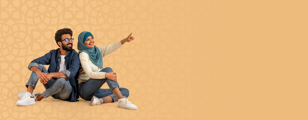 Two friends sit on a flat surface. One friend wears a hijab and points at something while the other...