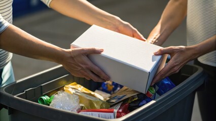 Obraz premium Two people placing a white box into a recycling bin filled with various recyclable materials like cans and plastic bottles.