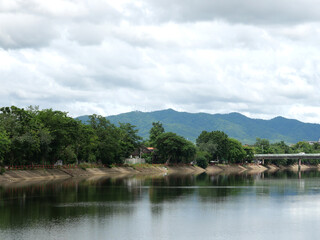 Fototapeta premium Calm river reflecting green trees and a bridge under a cloudy sky.