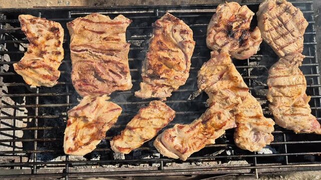 An overhead video of seasoned pork steaks with grill marks cooking on a metal charcoal barbecue grate over hot coals.