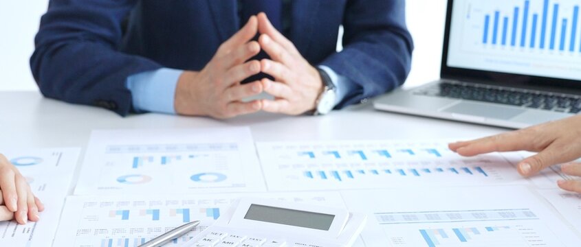 Business people reviewing financial charts and reports in office, using a laptop and calculator while engaging in discussions about strategy and performance