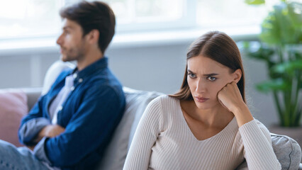 A man and a woman sit in a well-lit room with furniture and plants. The man appears distant while the woman shows signs of anger. They seem to be in a tense situation.