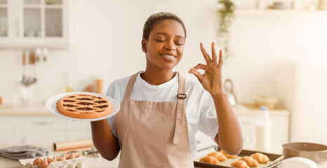 A woman stands in a bright kitchen holding a plate of cookies. She smiles and has a joyful expression while showing a hand gesture. The kitchen shows baked items in the background. © Prostock-studio