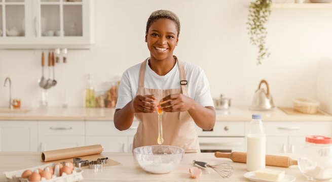 A woman stands in a well-lit kitchen, cracking an egg into a bowl while smiling. She wears an apron and is surrounded by baking tools and ingredients on the countertop.