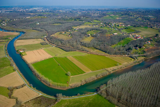 This captivating scene showcases the Nive River winding through lush fields and farmland near Villefranque. The diverse landscape features patches of green fields and rows of trees.