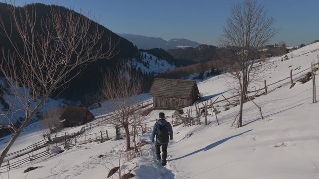 Cinematic tracking shot of a happy hiker walking down a snowy mountain trail among rustic barns at sunset, graded