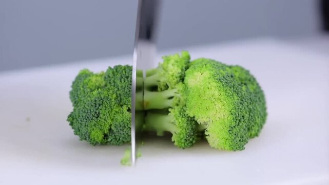 A knife slices through fresh green broccoli on a white cutting board, capturing the crisp texture and vibrant color as it cuts into smaller florets.