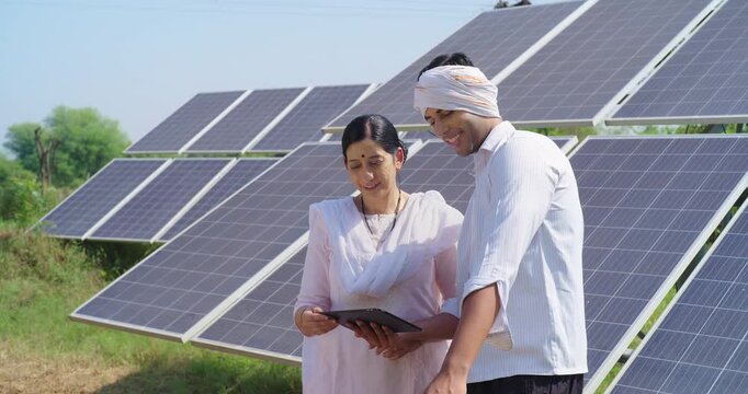 Indian mature adult woman hold tab look screen visit farm field do work check record data at rural area ground place female talk speak share eco friendly solar panel power plan idea to young male