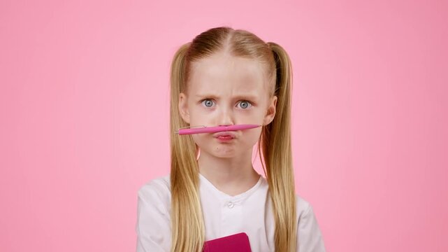 A girl with long blonde hair in two ponytails poses with a pen on her upper lip. She looks playfully serious in front of a bright pink background while holding a notebook.