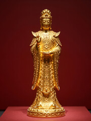 Fototapeta premium Elegant Golden Buddha Statue with Intricate Detailing and Warm Lighting Displayed on Red Pedestal Against Deep Maroon Background for Spiritual or Cultural Use
