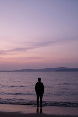 Fototapeta premium Silhouette of a Lone Person Standing at the Shoreline during Twilight with Calm Ocean Waves and Soft Pastel Sky in Background