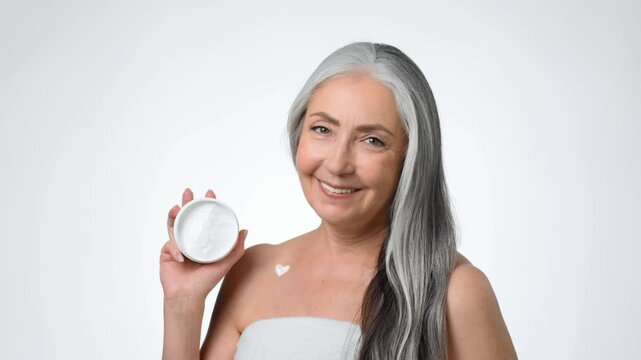 A woman with gray hair stands against a plain background. She holds a jar of cream in one hand and has a smile on her face. The scene conveys beauty and well-being.