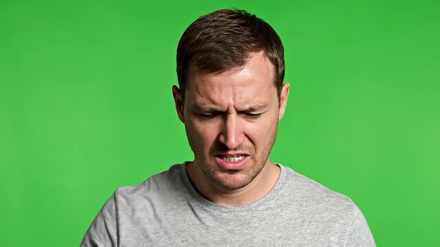A man with a frustrated expression looking down, standing against a green background, captured in a close-up shot
