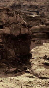 A large rock formation stands in a rugged area. The ground is covered with stones. No plants or animals are visible. The sun shines on the rocky surface.