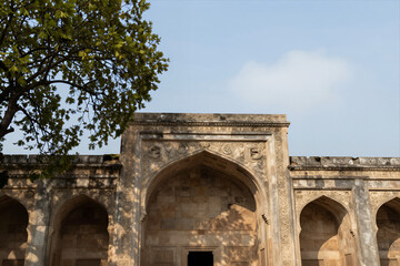 Historic Stone Mosque with Arched Entrance and Intricate Carvings under Clear Blue Sky Framed by Lush Green Tree Branches
