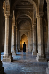 Solitary Figure in Dark Robe Walking Through Grand Arched Corridor of Ancient Stone Mosque with Intricate Carvings and Warm Ambient Lighting