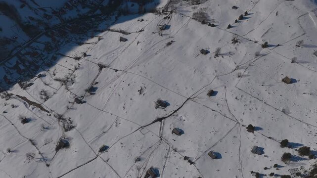 Top-down forward aerial shot over sunlit snowy hayfields revealing the shadowed village of Moieciu de Sus, graded