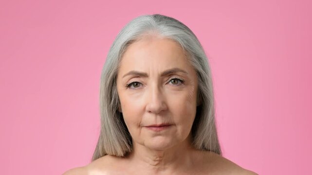 A mature woman with gray hair poses facing forward against a soft pink background. She exhibits a confident expression, showcasing her natural beauty and strength.