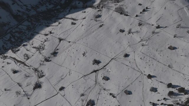 Top-down forward aerial shot over sunlit snowy hayfields revealing the shadowed village of Moieciu de Sus, ungraded log