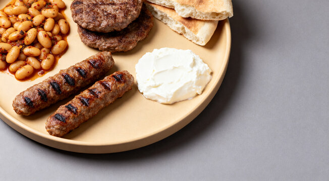 Authentic Balkan cuisine platter with cevapcici, pljeskavicas, kajmak, prebranac beans and pitas on beige tray over grey background.