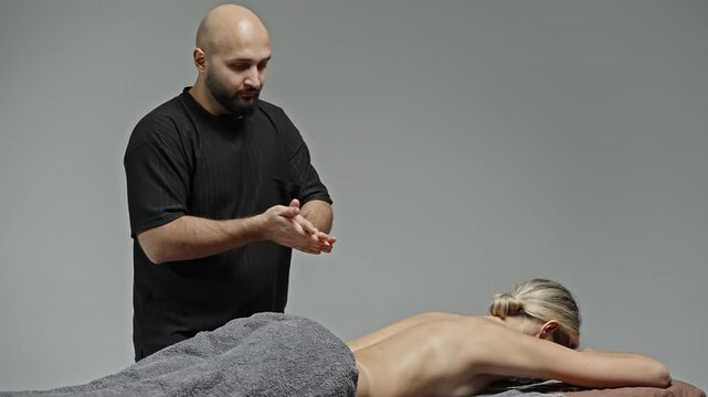 Professional massage therapy session as a therapist applies deep tissue techniques to a client's upper back while she lies face-down under a gray towel in a serene studio setting