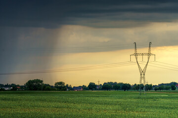 Słup energetyczny na zielonym polu pod dramatycznym, burzowym niebem. Widoczny opad deszczu w oddali i złote światło zachodu. Infrastruktura przesyłowa w wiejskim krajobrazie. © Henryk Niestrój
