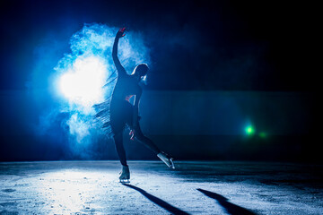 Silhouette of a figure skater performing on a dark indoor ice rink with bright blue backlight and...