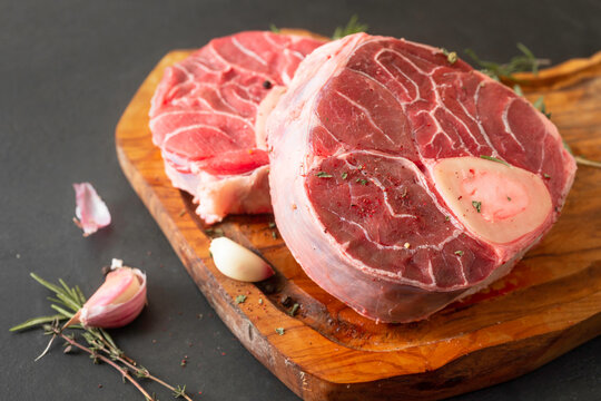Raw osso buco cuts with bone, placed on a rustic wooden cutting board, accompanied by fresh rosemary, thyme, and garlic cloves.