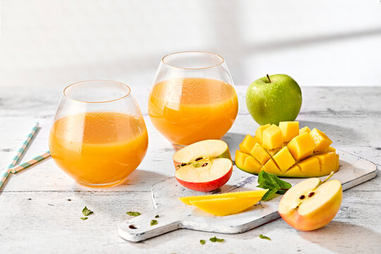 Apple and Mango Juice in Short Round Tumblers with Fresh Apples and Mango on White Wooden Surface
