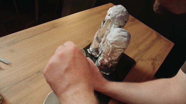 High angle view of caucasian pastry artist that applying icing on top of cupcake in foil wrap on wooden table. Bowl of glaze at side. Patient spooning of male chef in workspace. Culinary activities