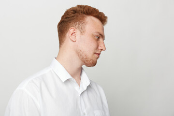 Profile of a young redhead man in a white shirt looking down with a neutral expression on a light grey background. Concept of contemplation, calmness, and casual style portrait. © SHOTPRIME STUDIO