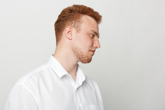 Profile of a young redhead man in a white shirt looking down with a neutral expression on a light grey background. Concept of contemplation, calmness, and casual style portrait.