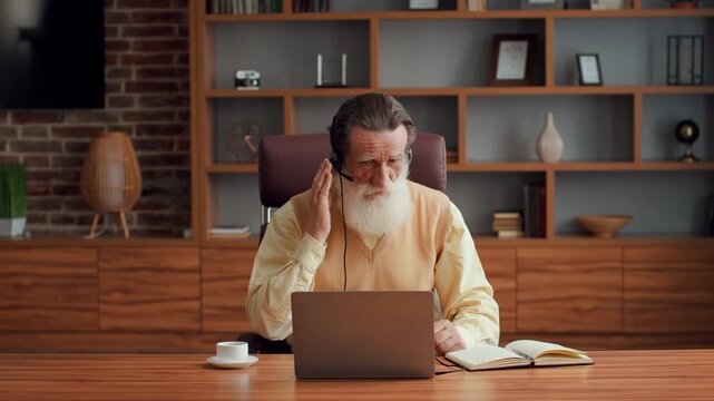 Education in any age. Bearded senior man wearing headset video chatting with tutor, writing notes in copybook, studying online at home, slow motion, free space