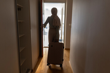 Fototapeta premium Muslim woman pushing suitcase down hallway, leaving home, standing near open door and an uncertain future filled with fear and sadness