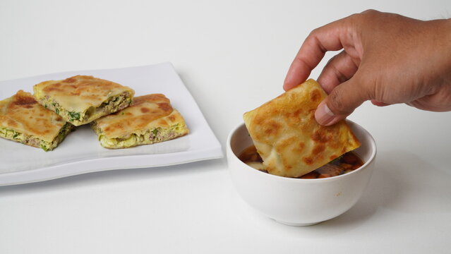 Hand holding martabak telur or martabak kubang served on a white plate with a bowl of pickled vegetables. This popular Indonesian street food features crispy fried stuffed pancake filled with egg and 