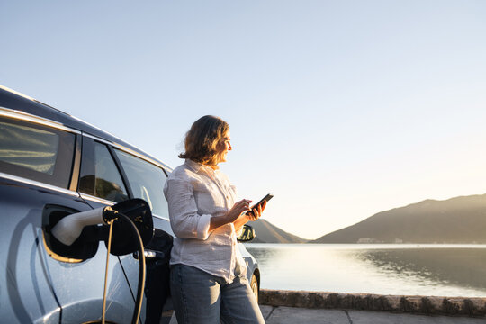Woman with smartphone next to charging electric car on the seacost during summer vacation