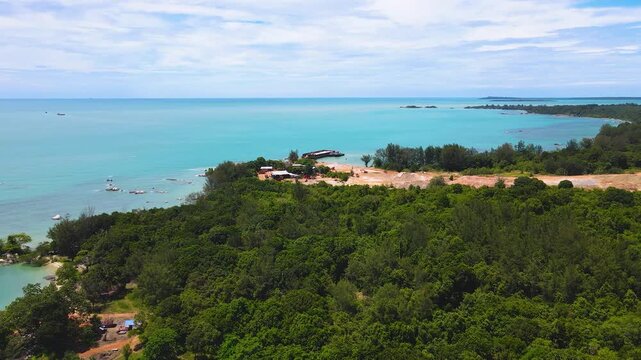 Breathtaking drone shot of Membalong's tropical coastline in Bangka Belitung, Indonesia. A pristine landscape of cyan sea, sandy beach, lush green trees, and mountains under a blue sky.