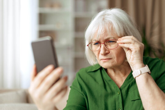 Senior woman adjusting eyeglasses while looking at smartphone screen. Mature caucasian female reading text or checking information on mobile device