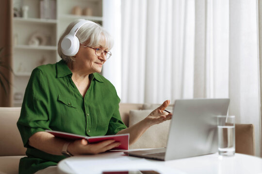 Senior woman wearing headphones and holding notebook while communicating through laptop at home. Mature caucasian female studying or attending online class in retirement lifestyle