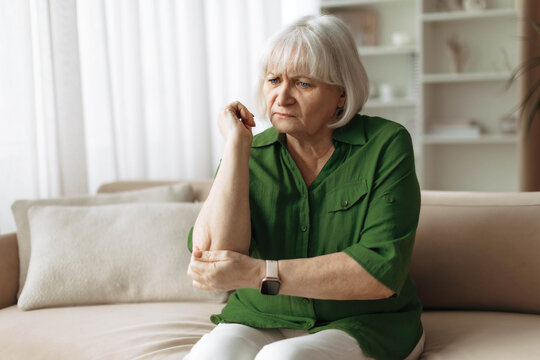 Senior woman touching painful elbow while sitting on sofa at home. Mature caucasian female experiencing joint pain or arthritis, showing health problems related to aging and retirement lifestyle