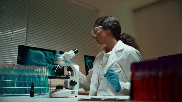 Young female microbiologist in a lab coat examining a test tube with blue liquid and taking notes. A microscope and DNA helix on a screen in a science lab